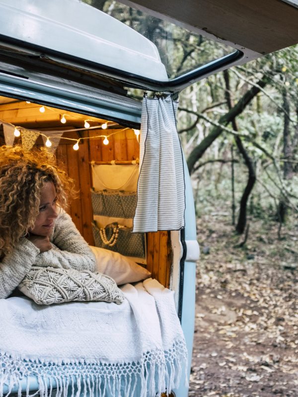 Happy free beautiful woman lay down and relax inside her blue van vehicle parked in the forest - people enjoying alternative travel and vacation or life options - female enjoying the camper van vacation